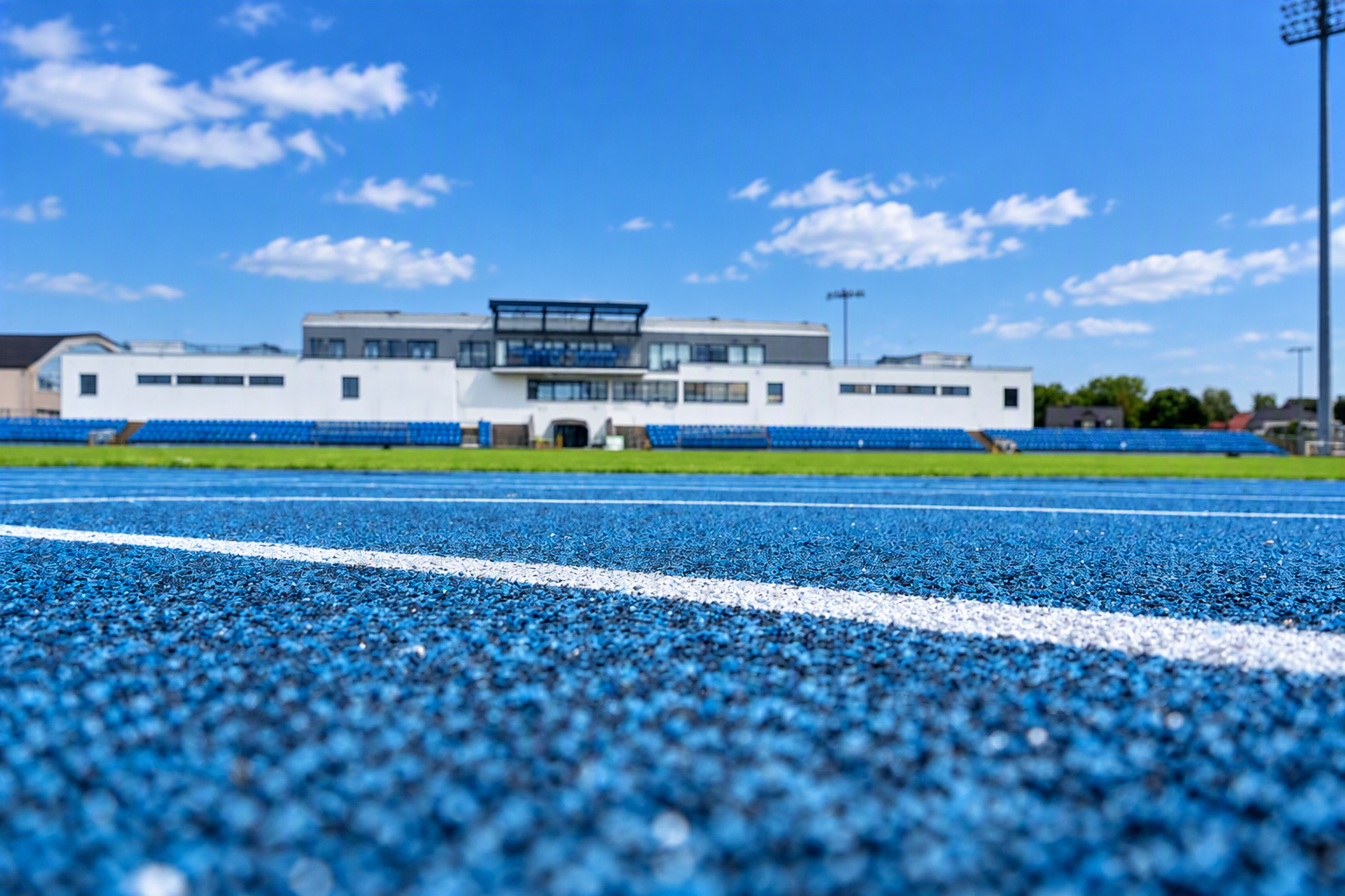 Widok stadionu z niebieską bieżnią lekkoatletyczną, boiskiem, trybunami i budynkiem zaplecza sportowego.