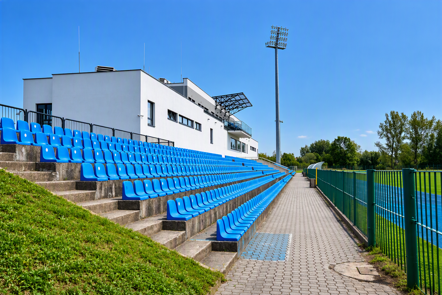 Trybuny stadionu z niebieskimi siedziskami, budynkiem zaplecza sportowego oraz masztem oświetleniowym.