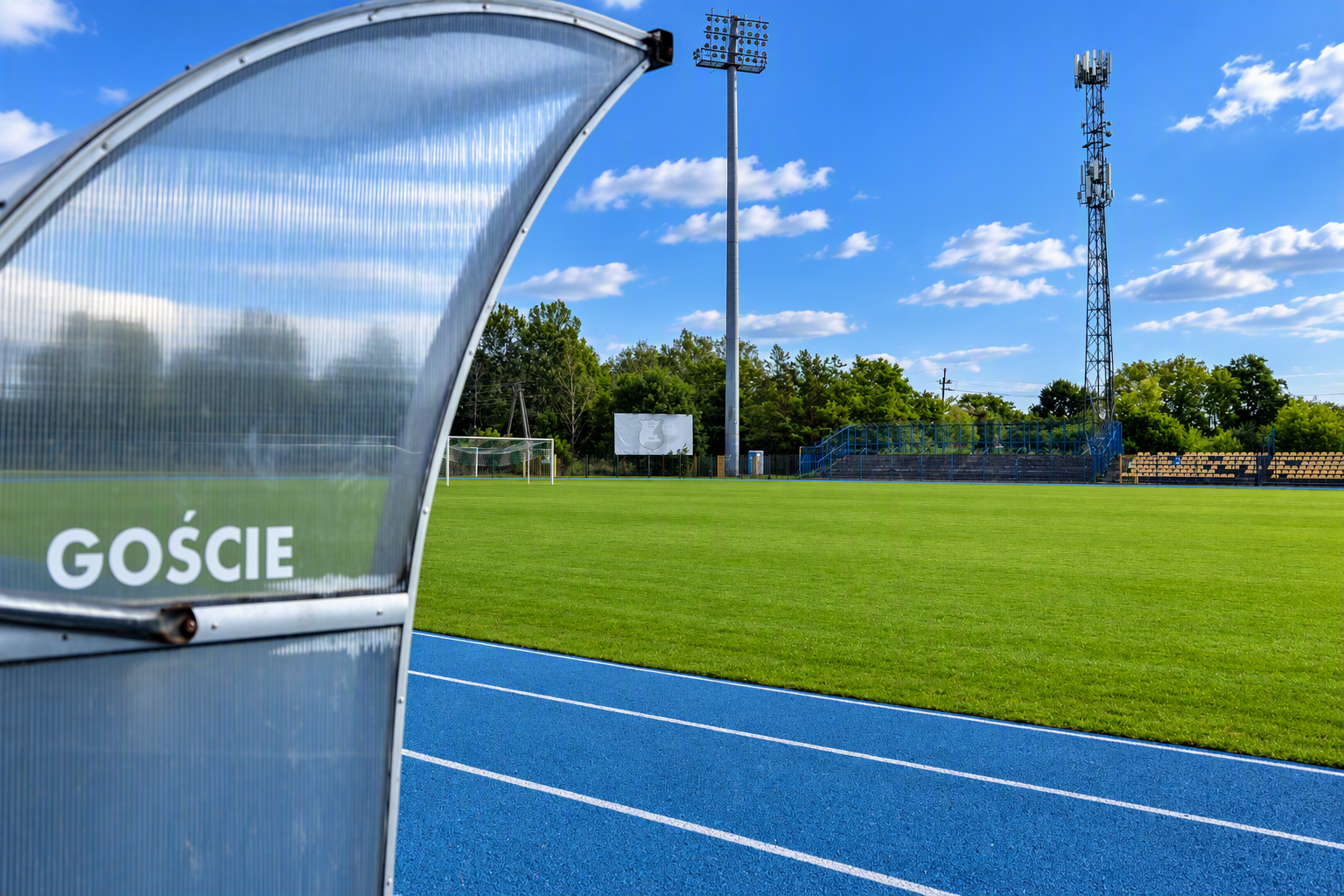 Boisko piłkarskie stadionu z niebieską bieżnią lekkoatletyczną, trybunami i ławką dla gości.